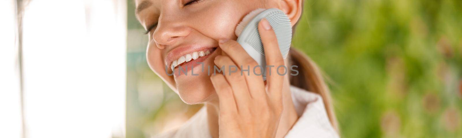 Cropped portrait of young woman wearing bathrobe smiling while using silicone face brush to cleanse skin by Yaroslav_astakhov