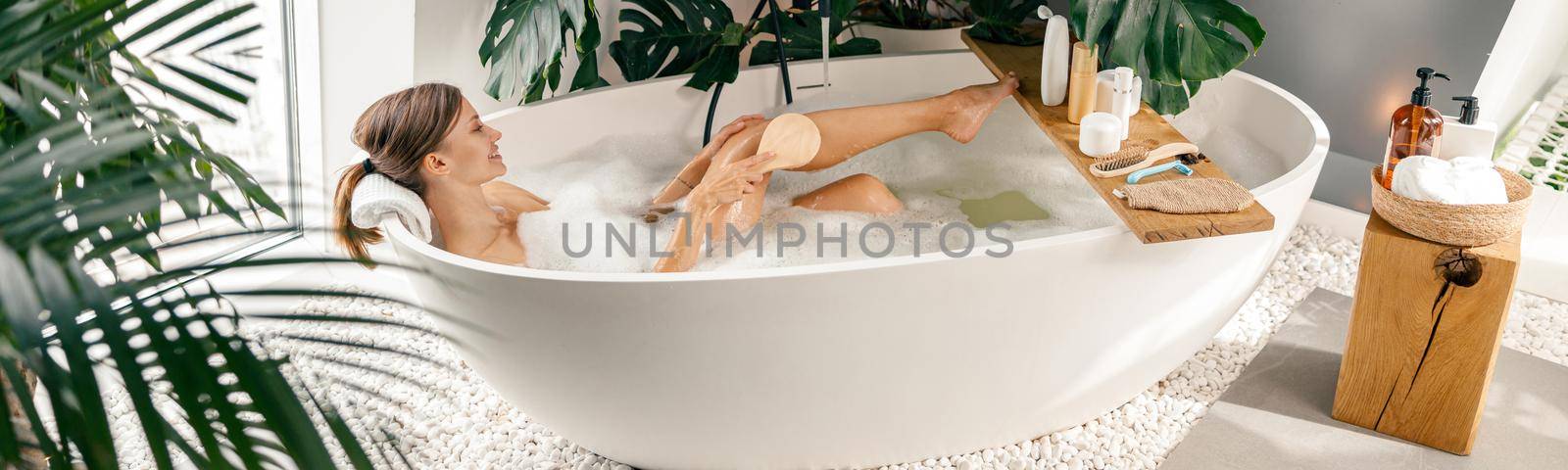 Relaxed young woman using brush while taking buble bath in modern bathroom decorated with tropical plants. Spa, beauty and care concept