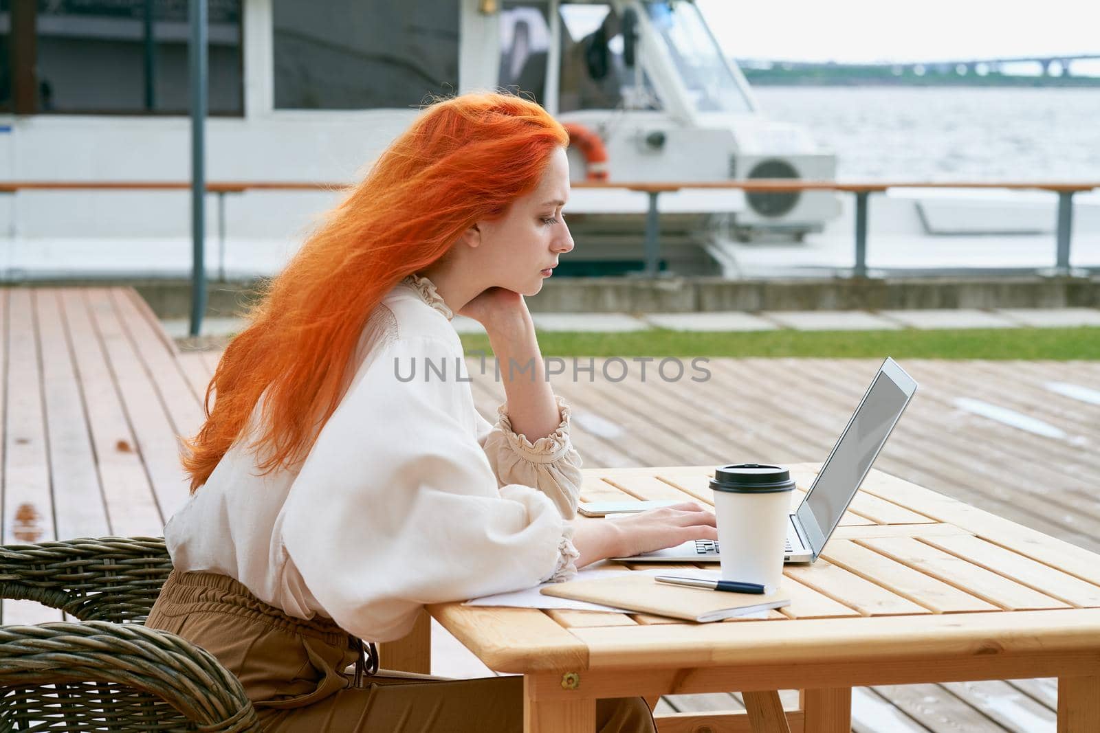Freelancer girl is sitting in cafe on street and remotely working on laptop. Redhaired businesswoman traveling, browsing Internet at seaport