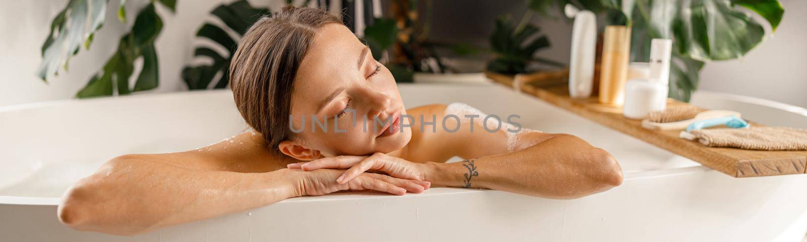 Dreamy young woman leaning on a bathtub side, relaxing with eyes closed at home by Yaroslav_astakhov