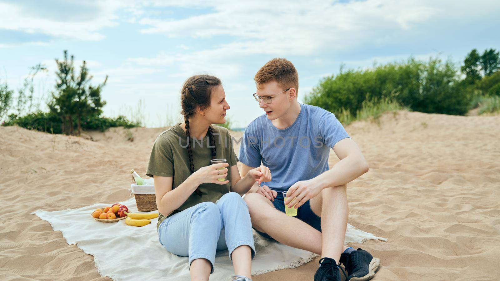 Boyfriend and girlfriend holding plastic cups full of beverage talking to each other smiling and sitting on blanket seashore background