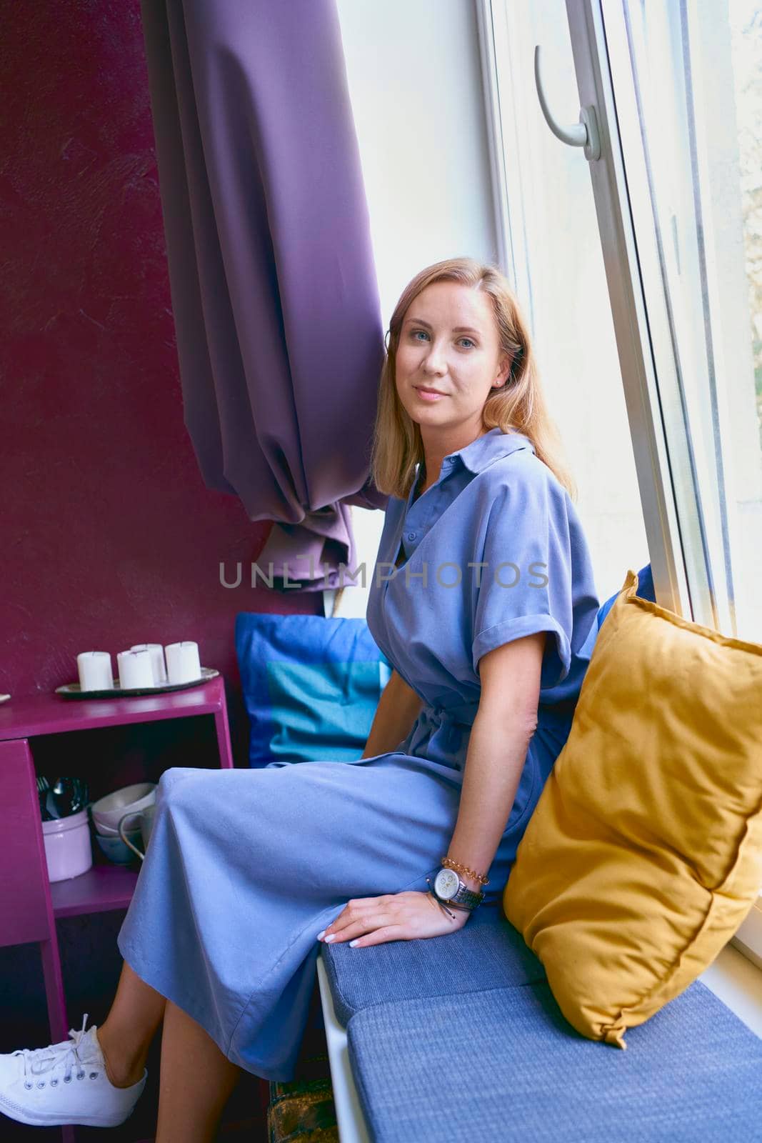 Beautiful self assured blonde woman with blue eyes in casual clothes dress and sneakers looking at camera while sitting on windowsill indoors