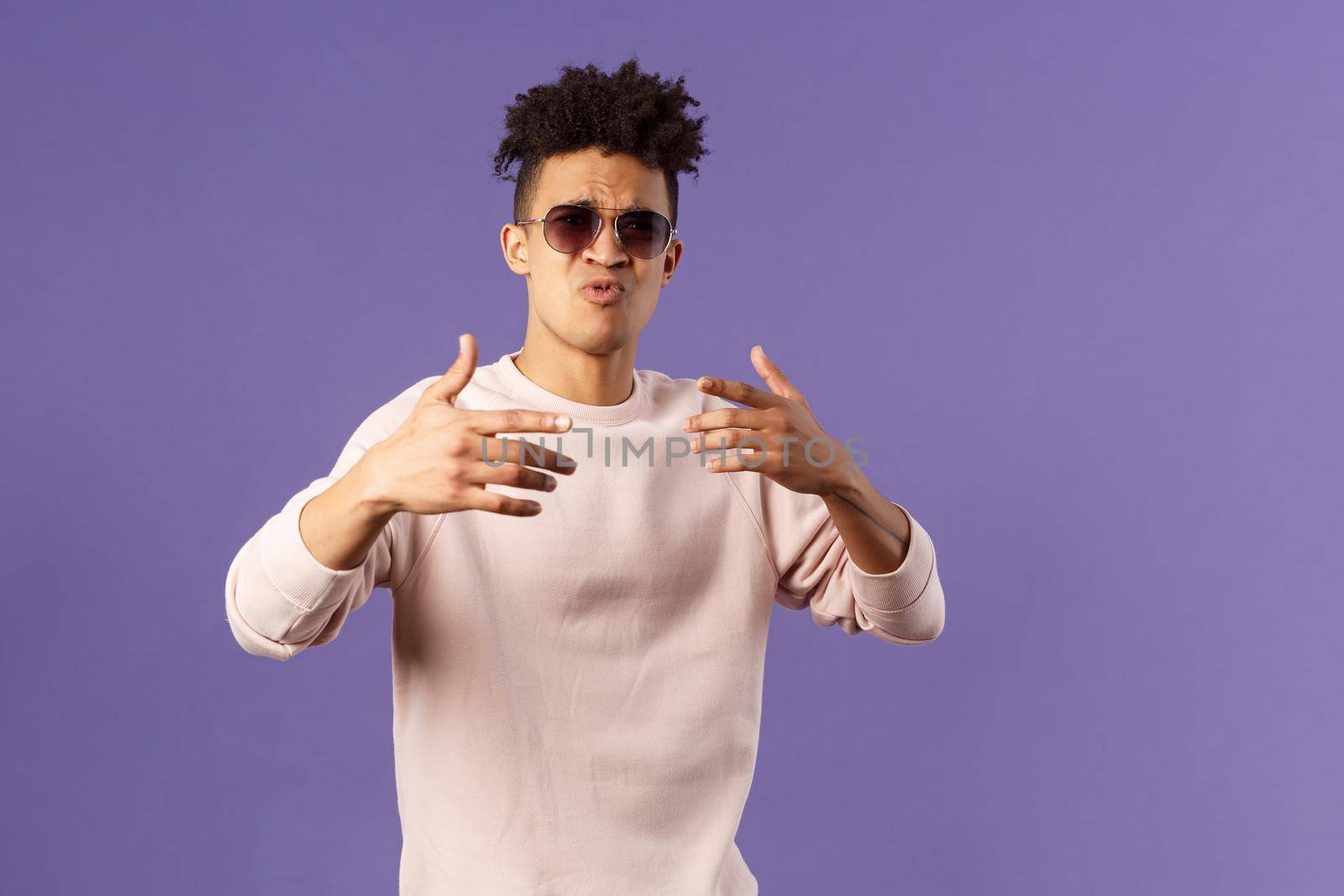 Portrait of sassy and carefree young hipster guy with dreads, wearing sunglasses dancing and gesturing while singing during rap battle, dance hip-hop on party, standing purple background.