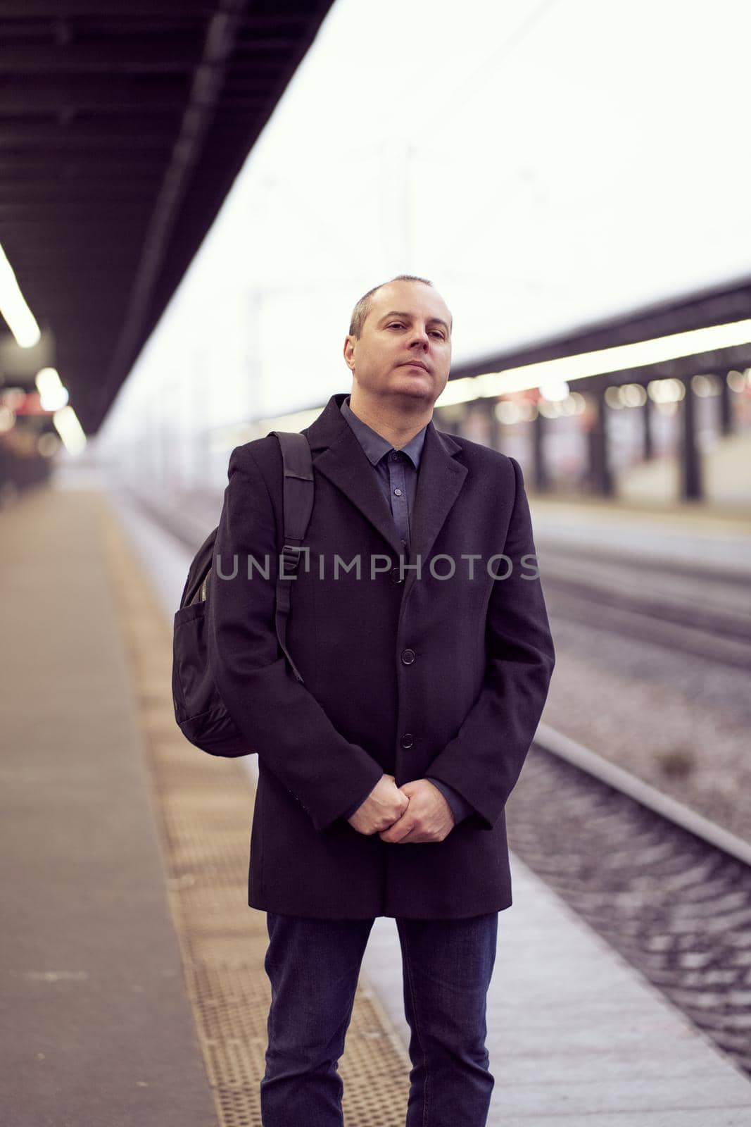 Train Station. A mature man in business clothes and a coat is standing and waiting for a train. Caucasian man looks into distance, winter or autumn. Dark evening. Vertical