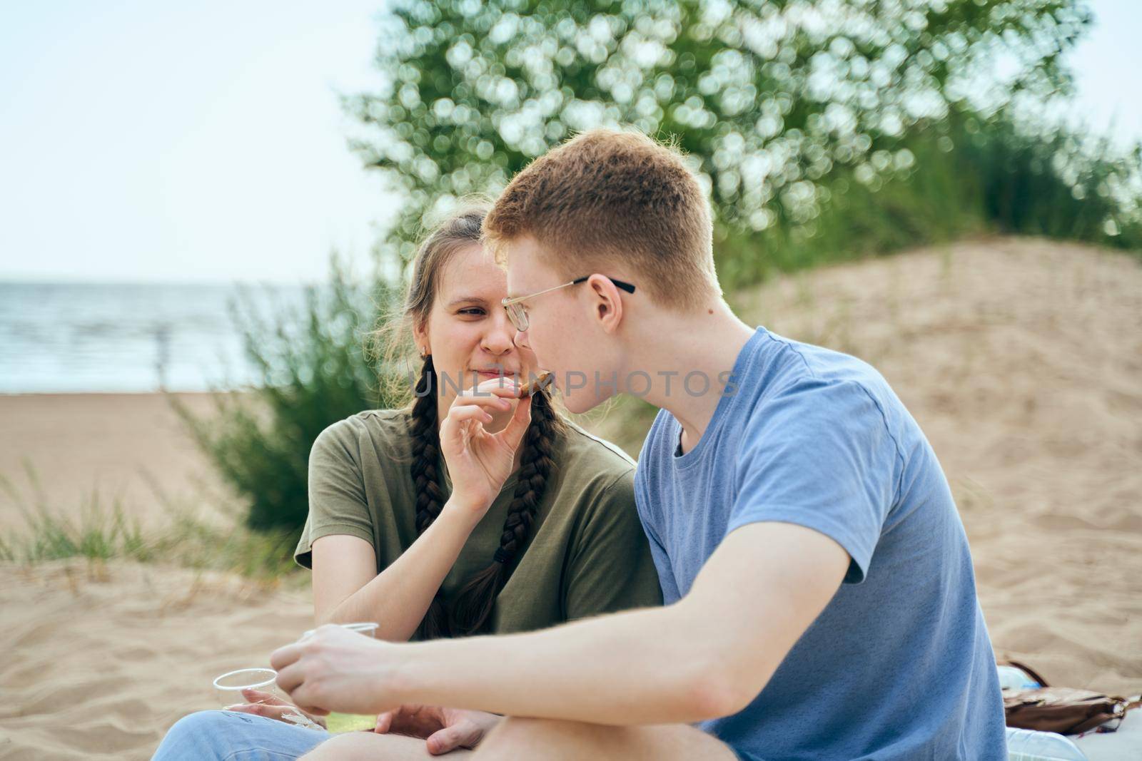 Side view of boyfriend and girlfriend feeding each other smiling and holding plastic cups during picnic on seaside background