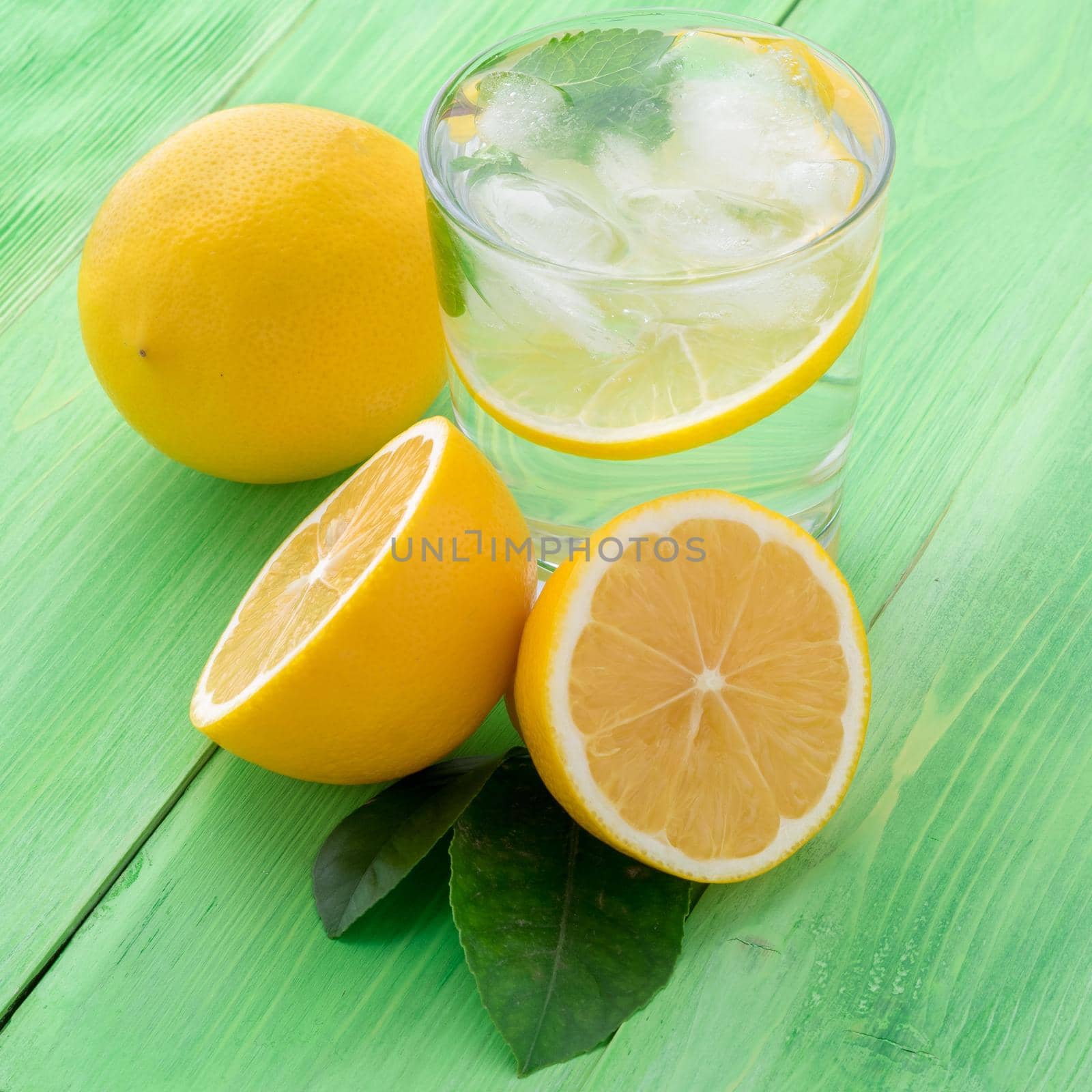 Lemonade in a glass, a lemon half, fresh leaves on the green table. A refreshing cold drink of water with ice, mint and slices of lemon, side view
