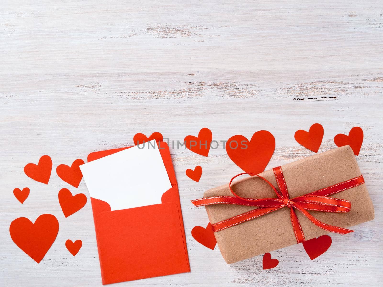 symbol of Valentine's day - gift box in kraft brown paper with red ribbon, envelope with a clean blank sheet of paper on white background with lots of red hearts, side view