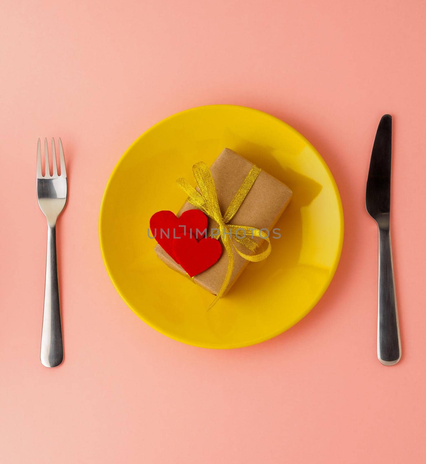 gift, pack of Kraft brown paper with a gold ribbon, two felt hearts on a yellow plate, knife, fork on a bright pink background, top view, copy space for text. Valentine's day . by NataBene