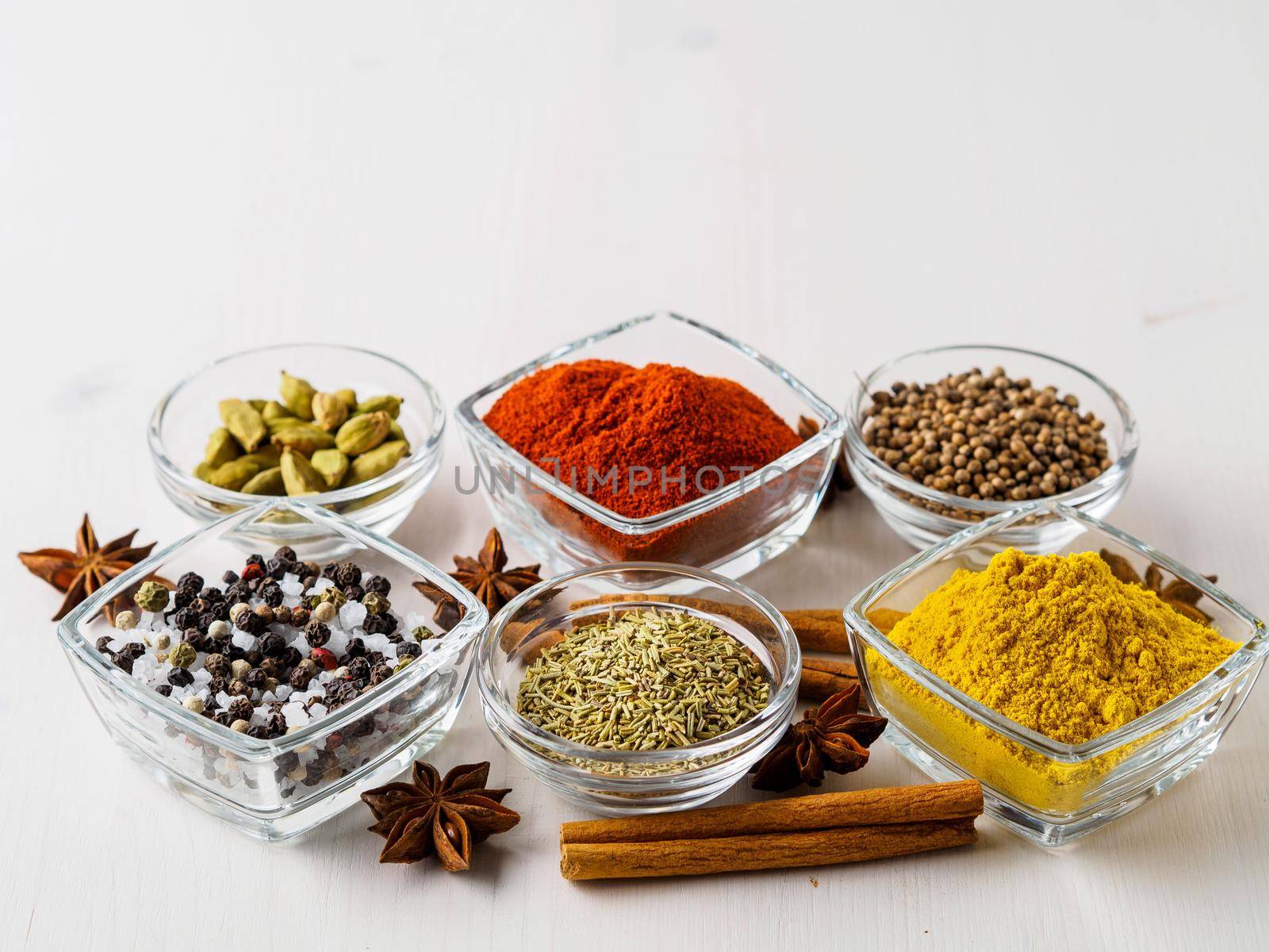 spice set-coriander, red pepper, turmeric, cinnamon, star anise, rosemary various seasonings in glass cups, on white wooden table, side view, blank space for text. by NataBene