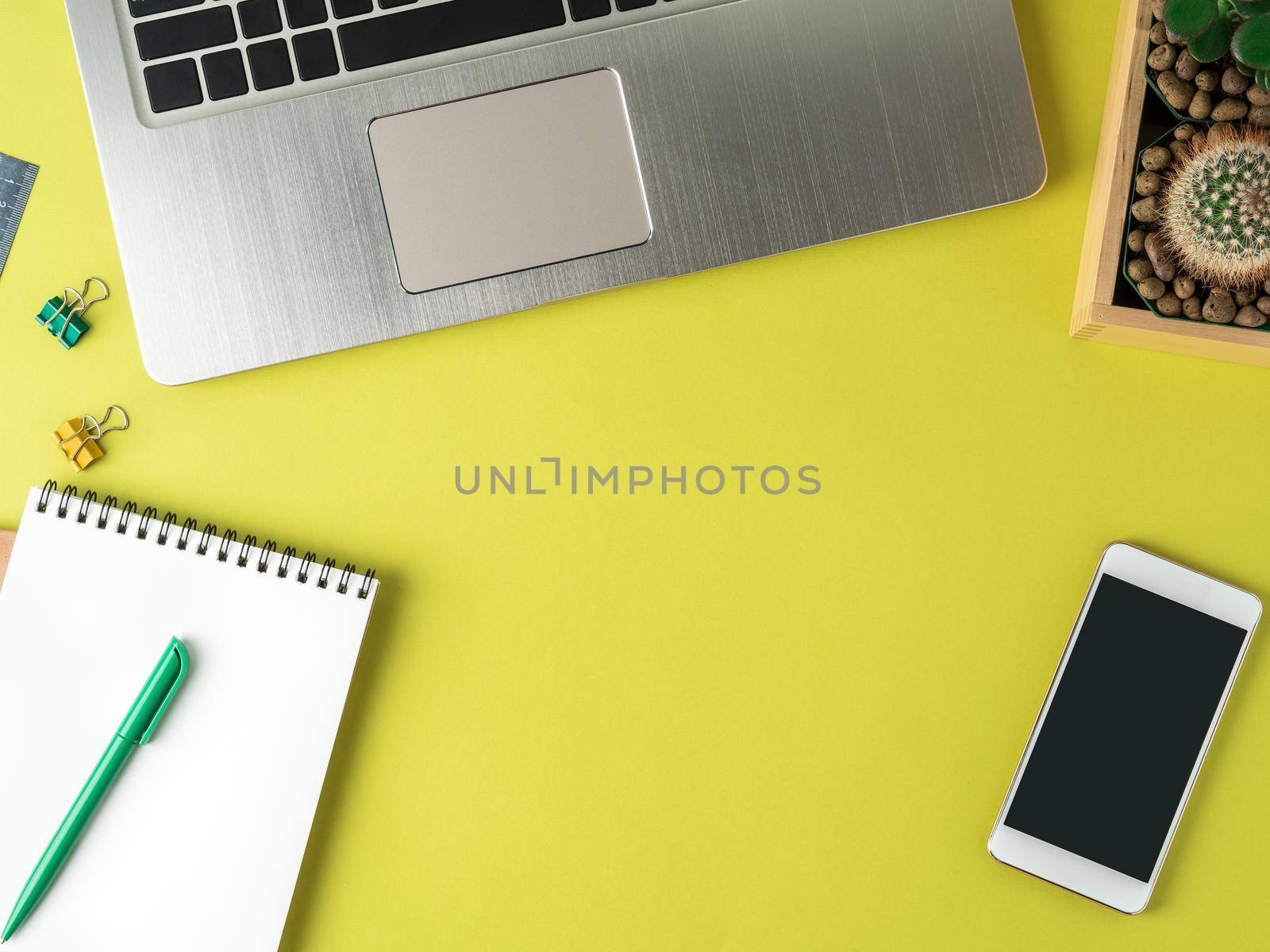 Top view of modern bright yellow office desktop with blank notepad, computer, smartphone, cactus, succulent. Mock up, empty space by NataBene