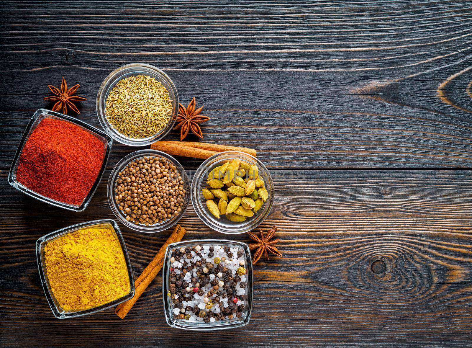 Set of spices on a dark brown textured wooden table - coriander seeds, ground red pepper, salt, black pepper, rosemary, turmeric, curry, coriander, star anise, cinnamon. Top view, empty space