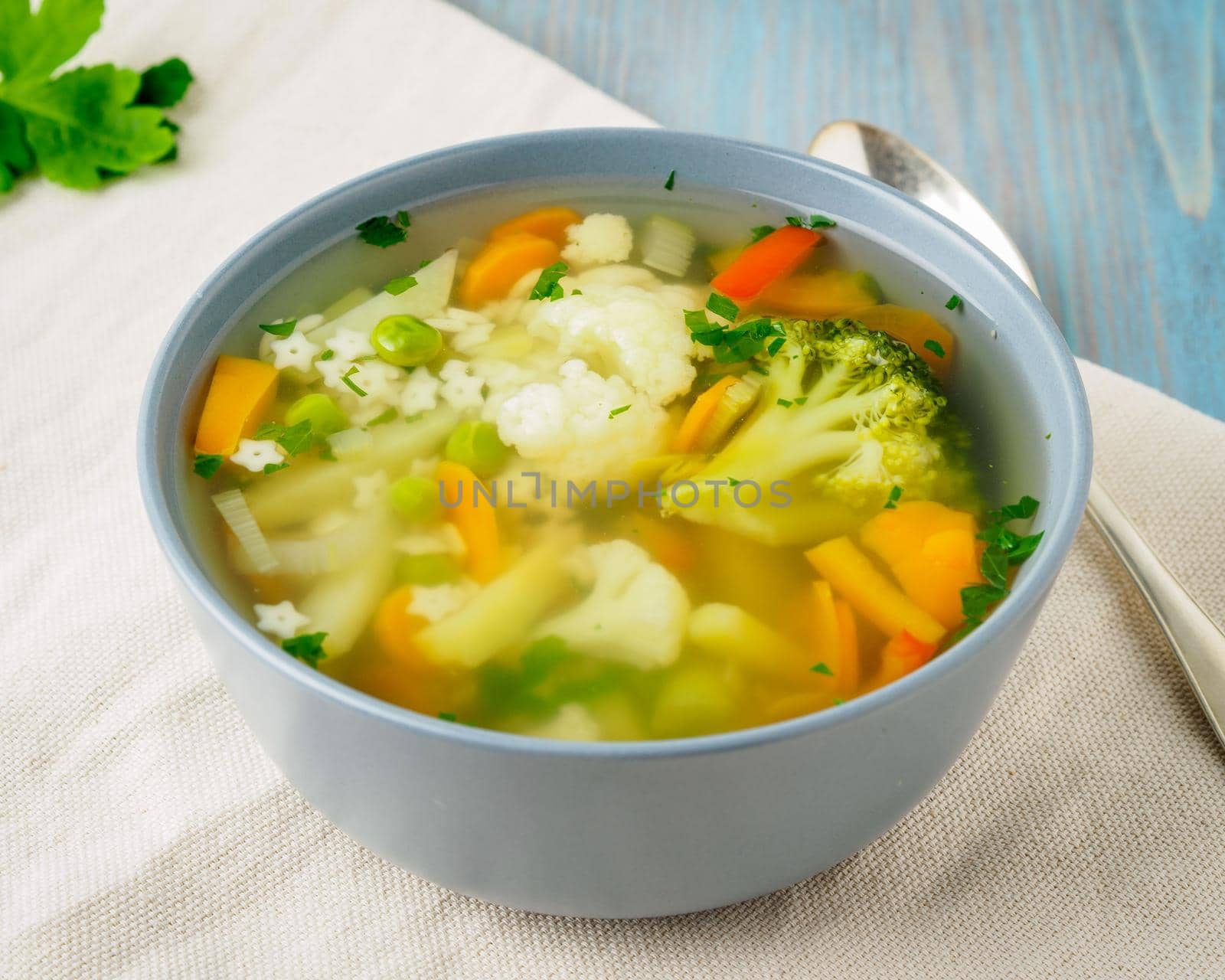 Bright spring vegetable dietary vegetarian soup with cauliflower, broccoli, pepper, carrot, green peas, pasta, parsley. Side view, blue background. by NataBene