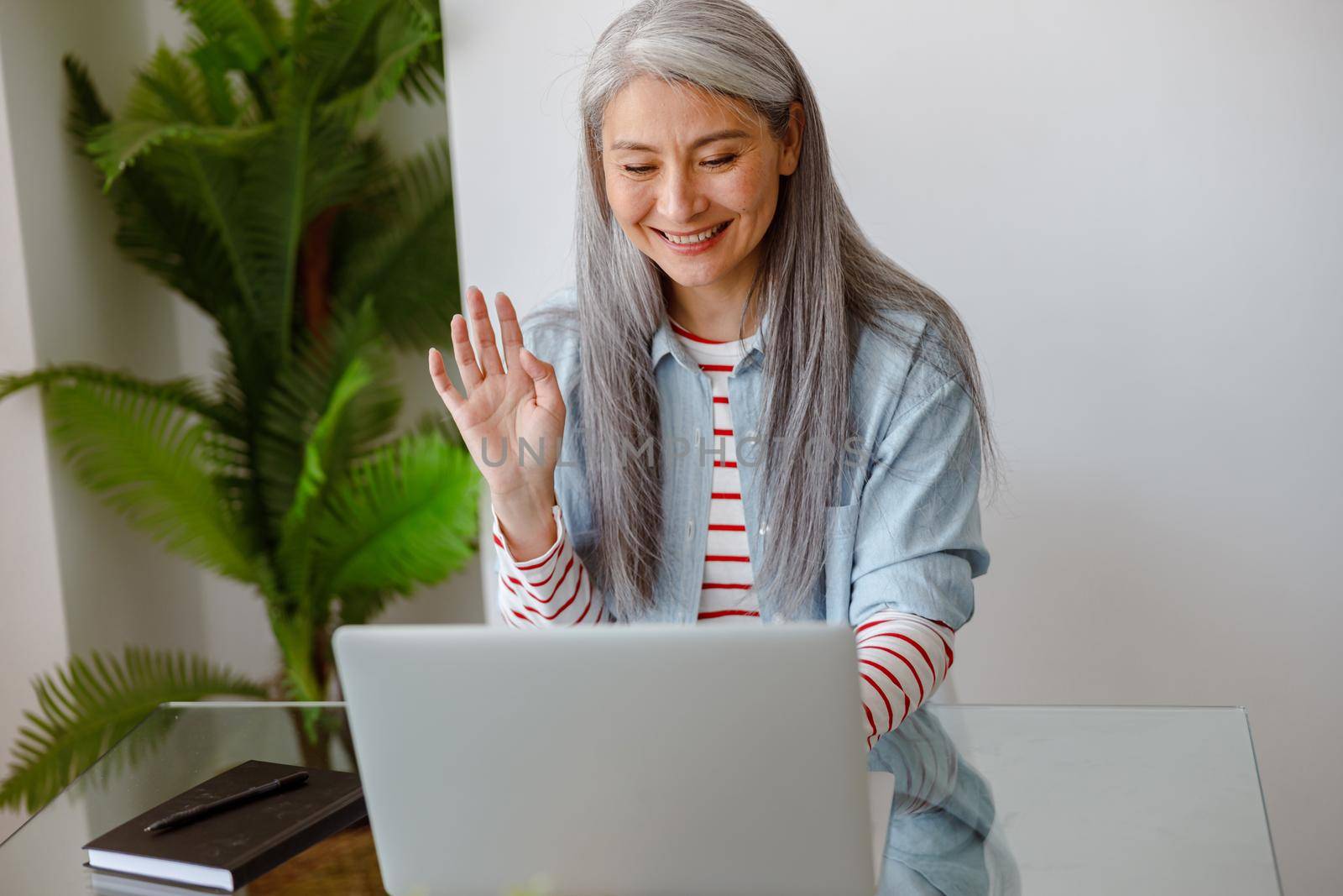 Joyful female person doing hello gesture and smiling while sitting at the table with notebook and talking with friend through video call