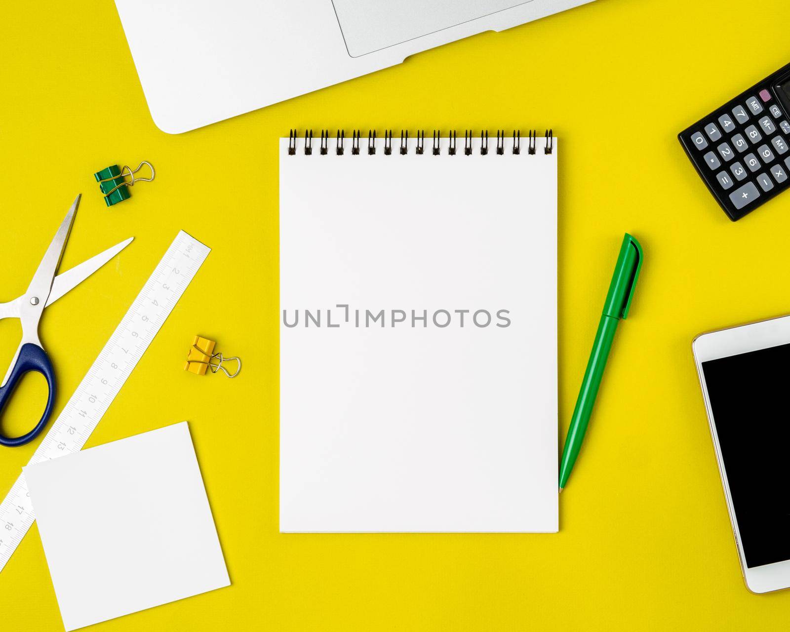 Modern creative bright yellow office desk with laptop, smartphone and other accessories - calculator, scissors, ruler, pen. Blank Notepad page for text in the middle, top view.. by NataBene