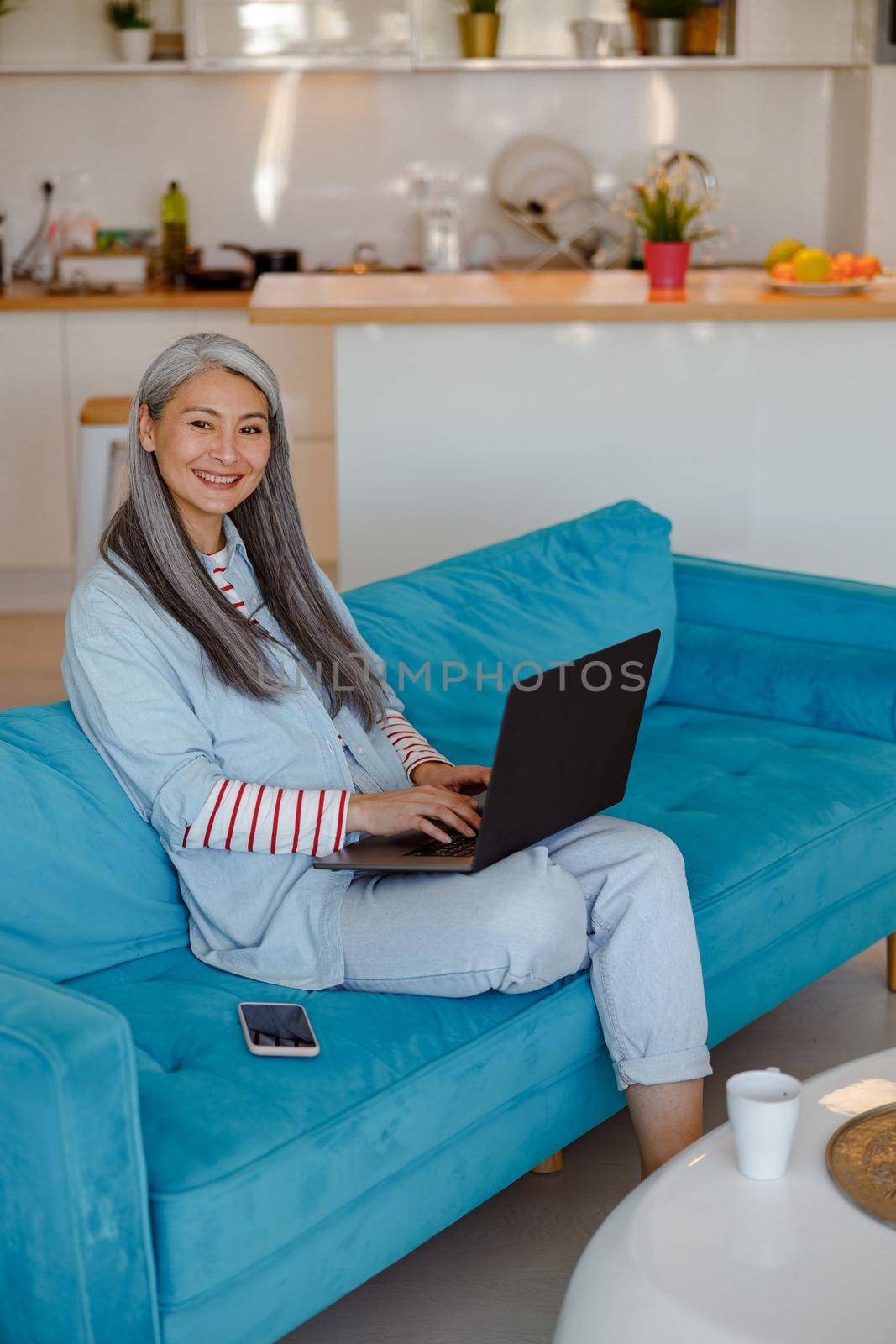 Joyful lady looking at camera and smiling while sitting on sofa and working on modern notebook