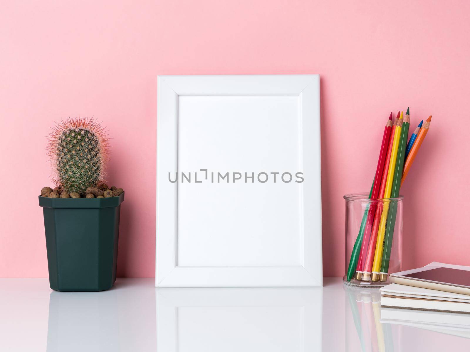 Blank white frame, crayon in jar, plant cactus on a white table against the pink wall with copy space