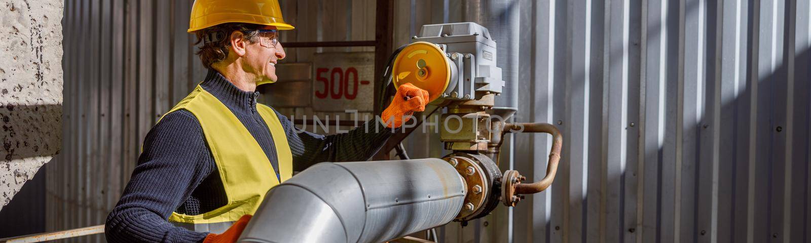 Cheerful matured man factory worker wearing work vest and gloves while standing near metal pipe and smiling