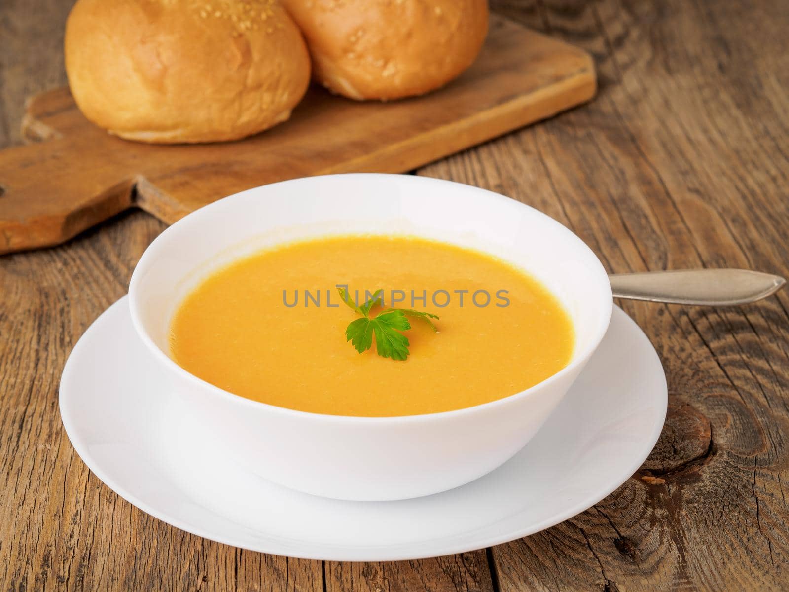 white bowl of pumpkin soup, garnished with parsley on wooden background, side view.