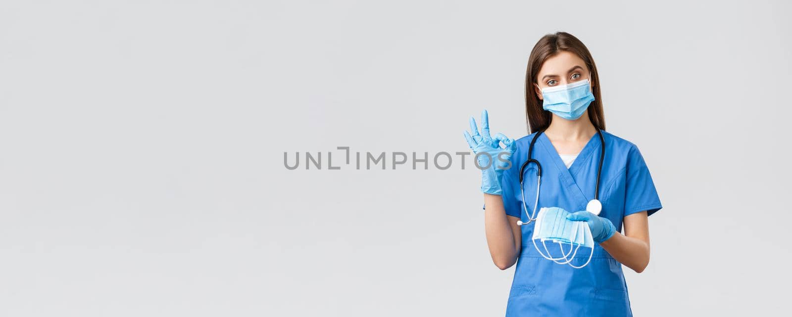 Covid-19, preventing virus, health, healthcare workers concept. Professional female nurse or doctor in blue scrubs and personal protective equipment, show okay sign and give medical mask by Benzoix