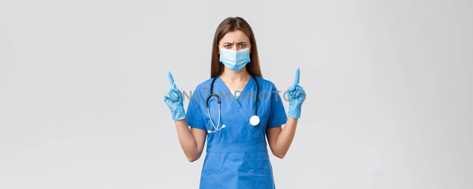 Covid-19, preventing virus, health, healthcare workers and quarantine concept. Skeptical and disappointed female nurse in blue scrubs feel displeased, pointing fingers up, wear medical mask by Benzoix