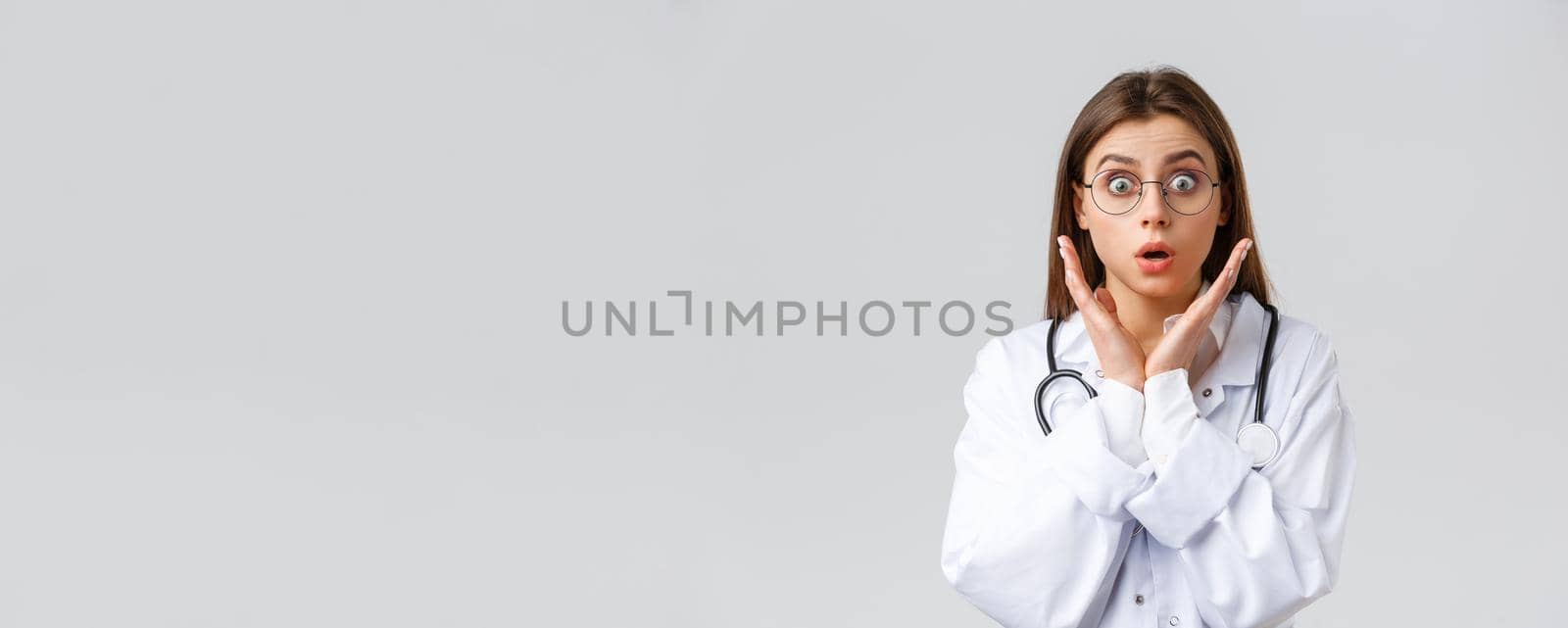 Healthcare workers, medicine, insurance and covid-19 pandemic concept. Surprised and impressed female doctor in glasses, white scrubs, gasping and staring astonished camera, grey background.