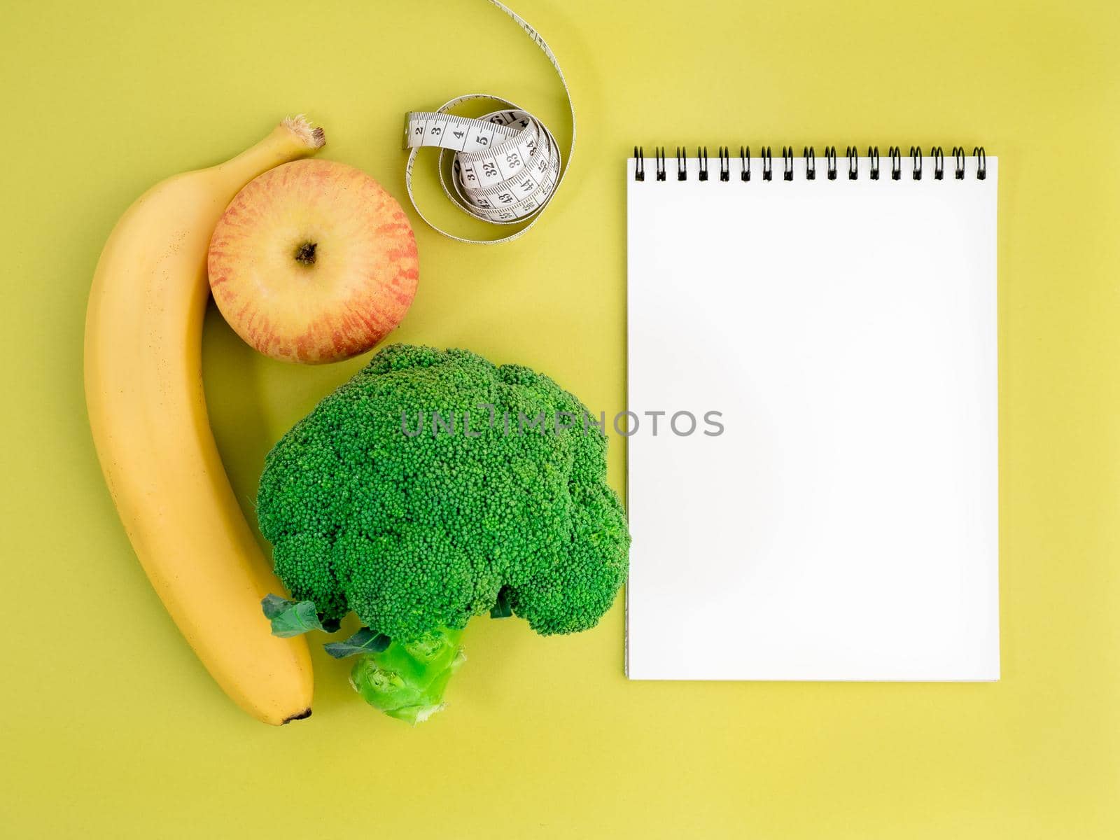 Fruits and vegetables - apple, banana and broccoli on bright yellow background. Notebook to record about diet.