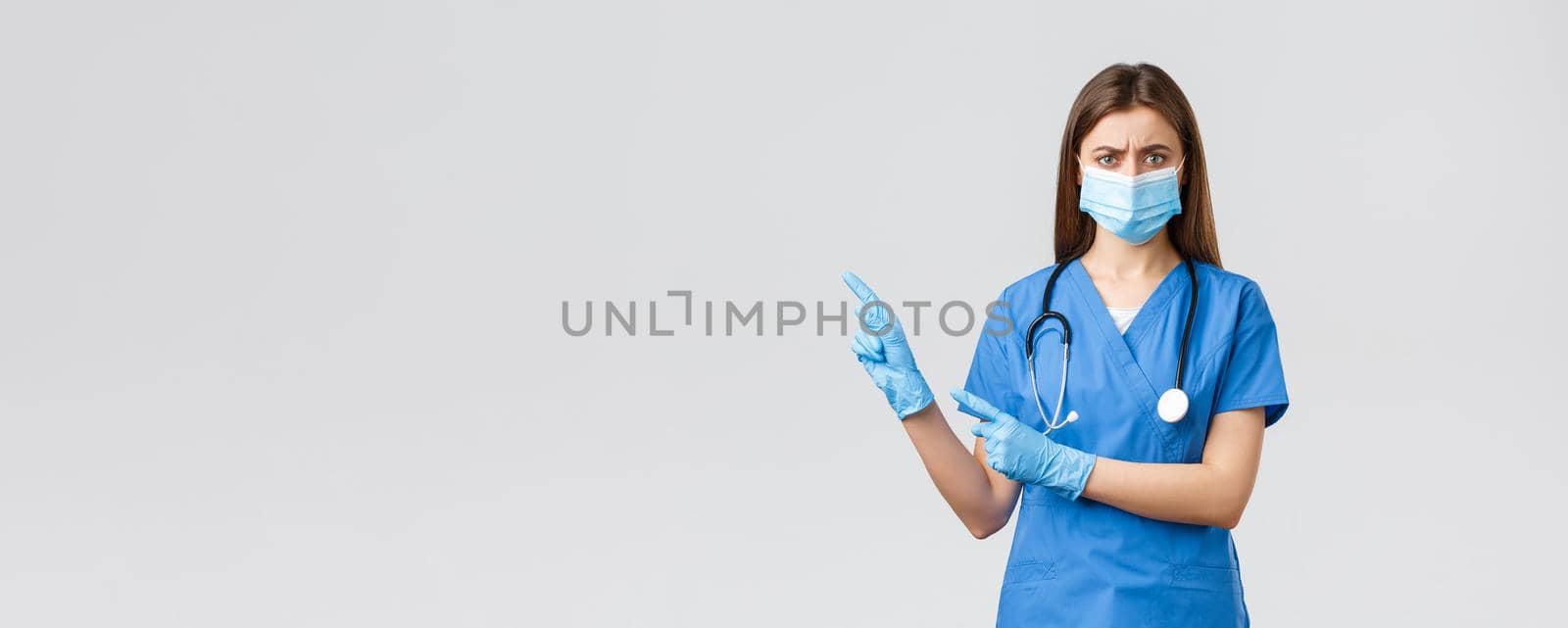 Covid-19, preventing virus, health, healthcare workers and quarantine concept. Angry concerned female nurse in blue scrubs, doctor wearing medical mask, frowning and pointing left at bad info.