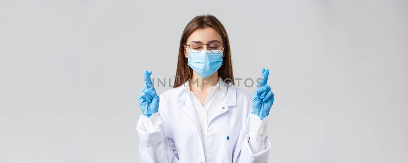 Covid-19, preventing virus, healthcare workers and quarantine concept. Hopeful doctor in white scrubs, medical face mask and gloves, close eyes, cross fingers and praying patients cure infection.