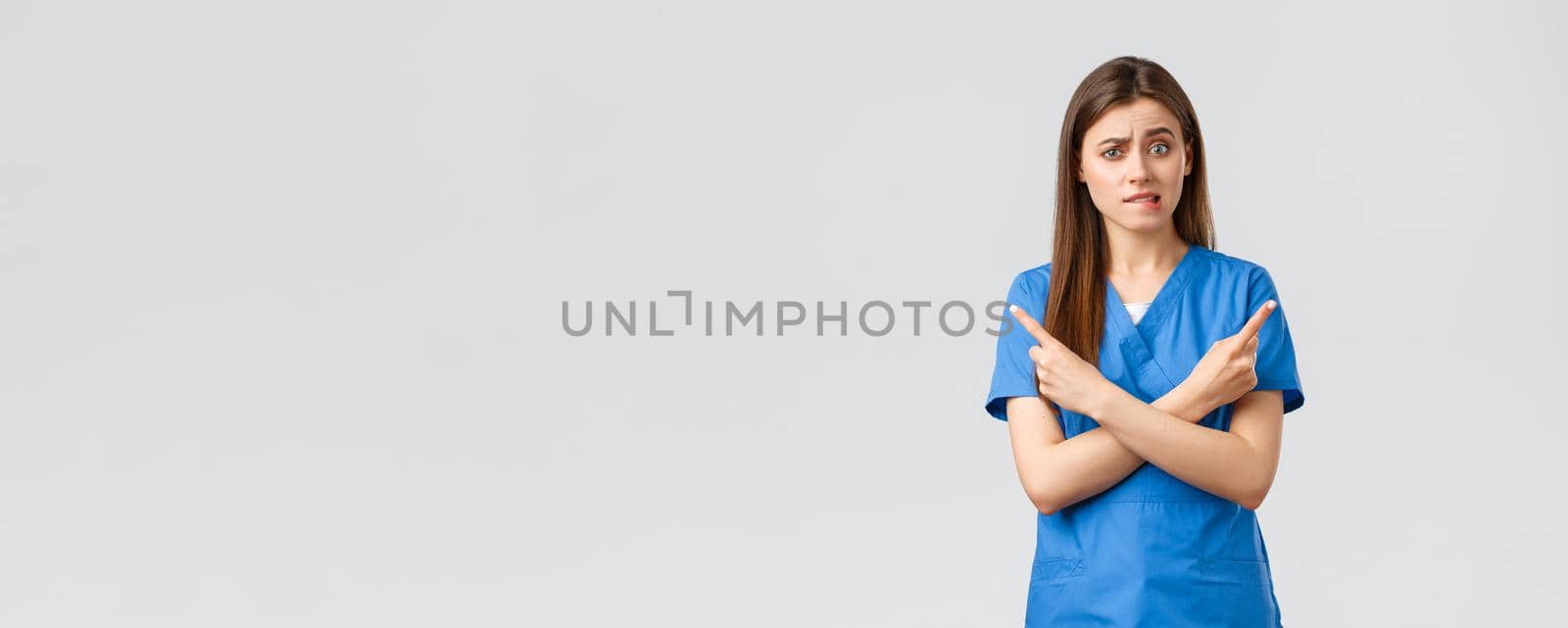 Healthcare workers, prevent virus, insurance and medicine concept. Indecisive and puzzled female nurse, doctor in blue scrubs pointing sideways at banners, biting lips nervous, need advice.