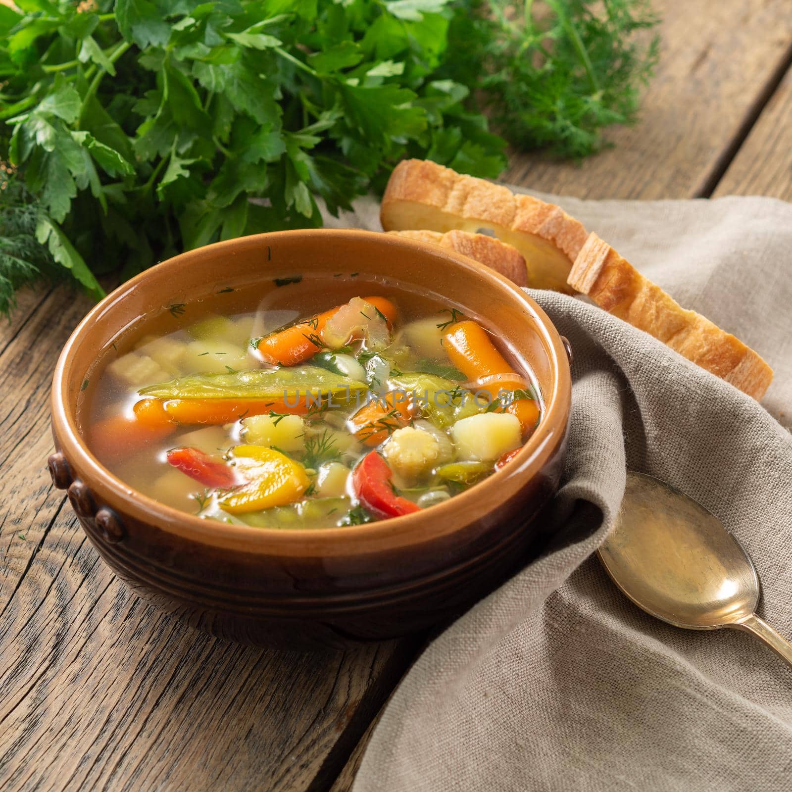 Bright spring vegetable dietary vegetarian soup with potatoes, pepper, carrot, green peas, parsley. Side view, brown rustic wood background, linen napkin.