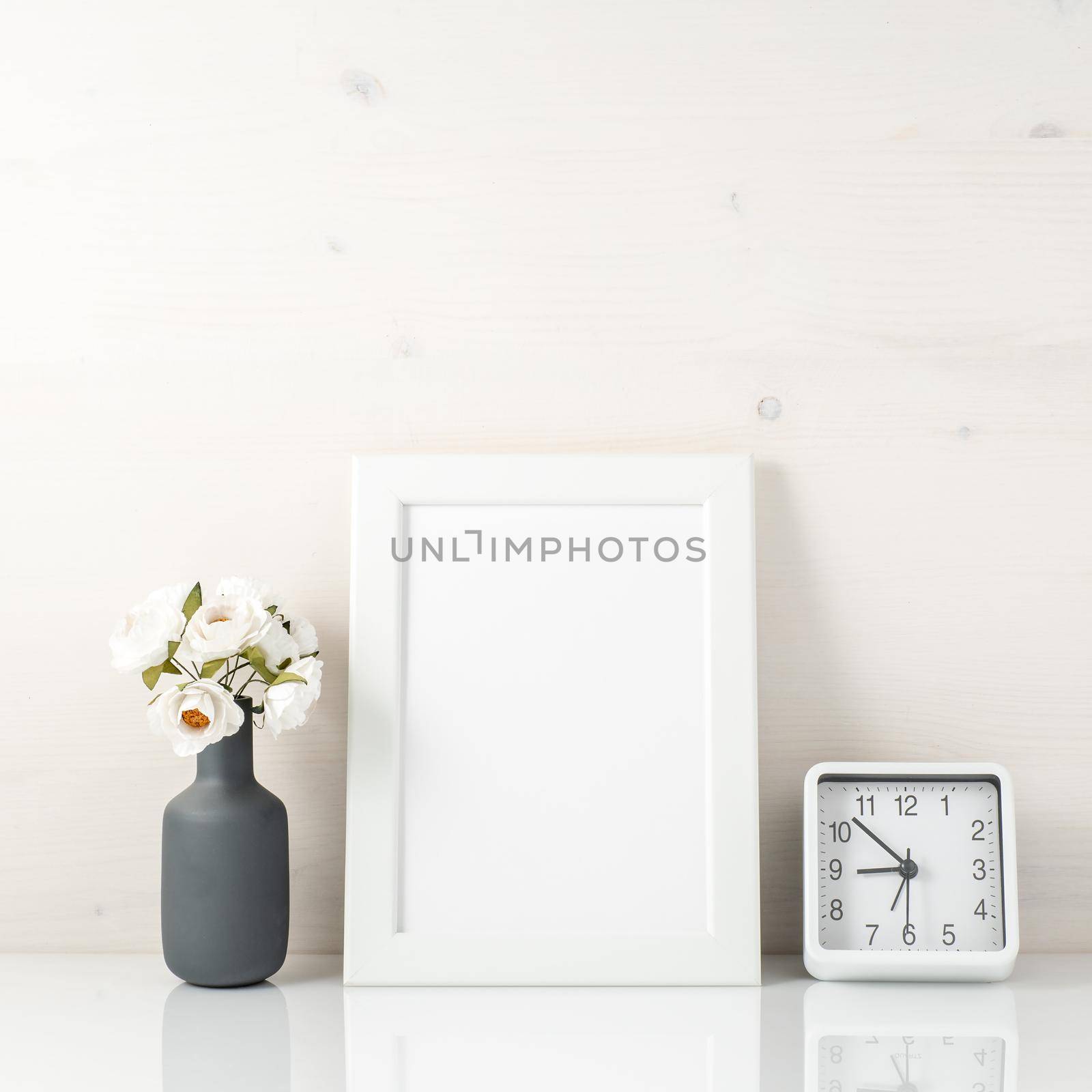 White frame, flower in vase, clock on a white table against the white wall with copy space. Mock up.