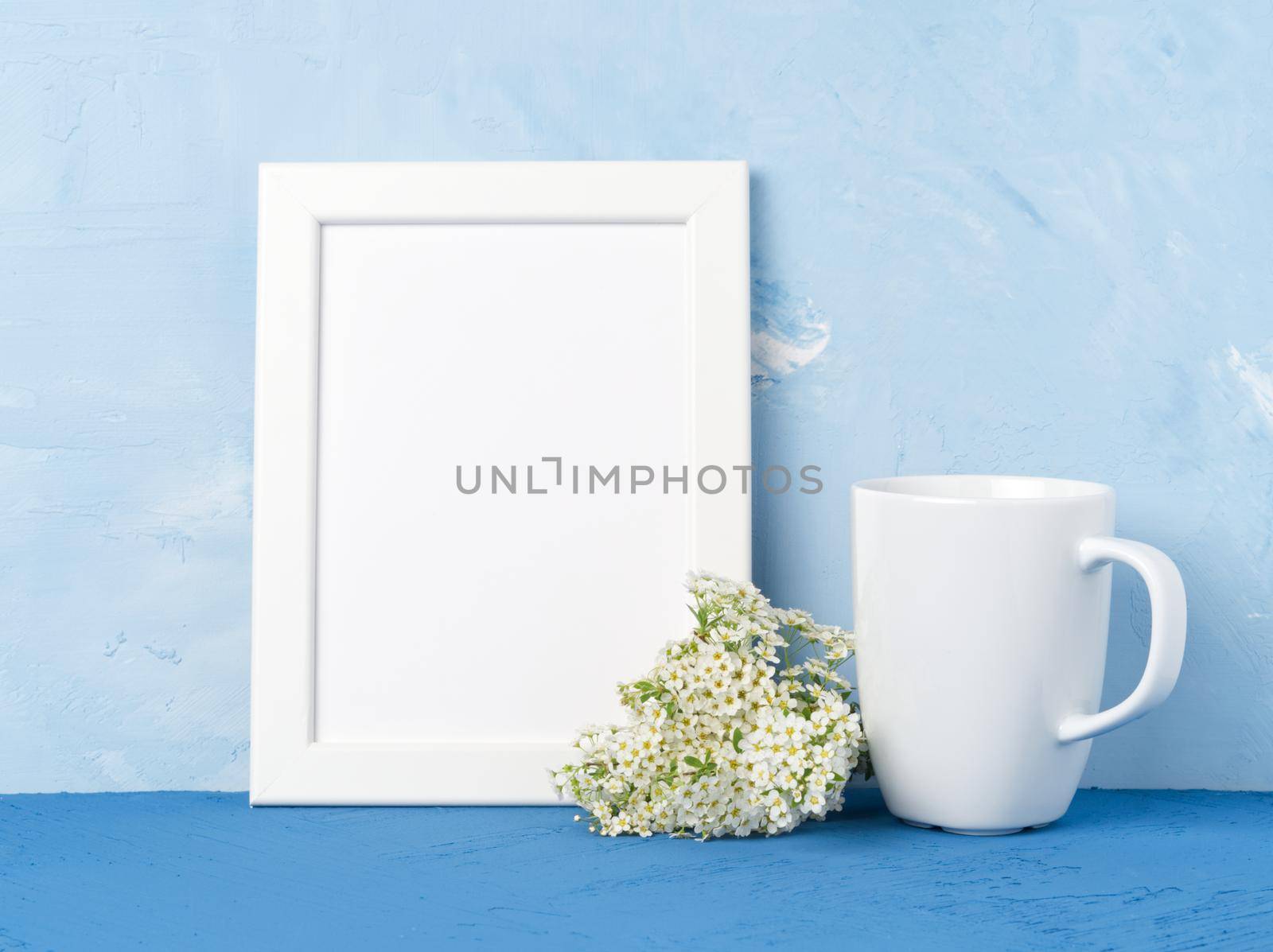 White mug with tea or coffee, frame, flower bouquet on the blue table opposite blue concrete wall. Mock up with empty space