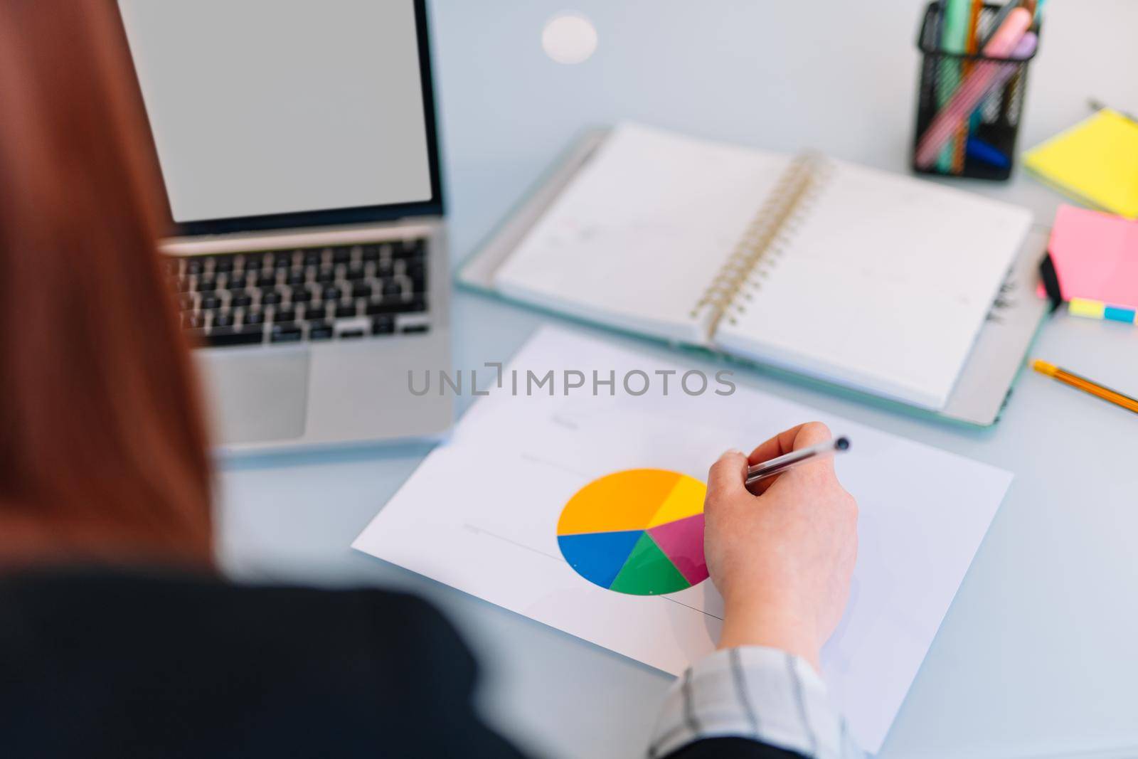 Young, red-haired business girl working on a chart in the office or at home while searching for information on the computer. Blank computer screen. She has a notebook with notes and a chart she is teleworking on. The girl is wearing jeans and a black blazer.