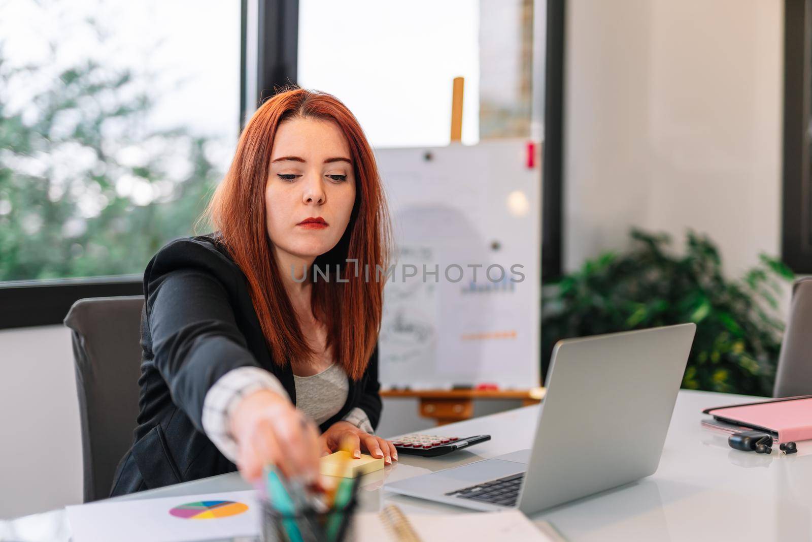 Pretty young redheaded pretty woman teleworking from home. She is making a work video call explaining something. Very bright environment with a large window behind and natural light. Young businesswoman and entrepreneur working on white laptop.