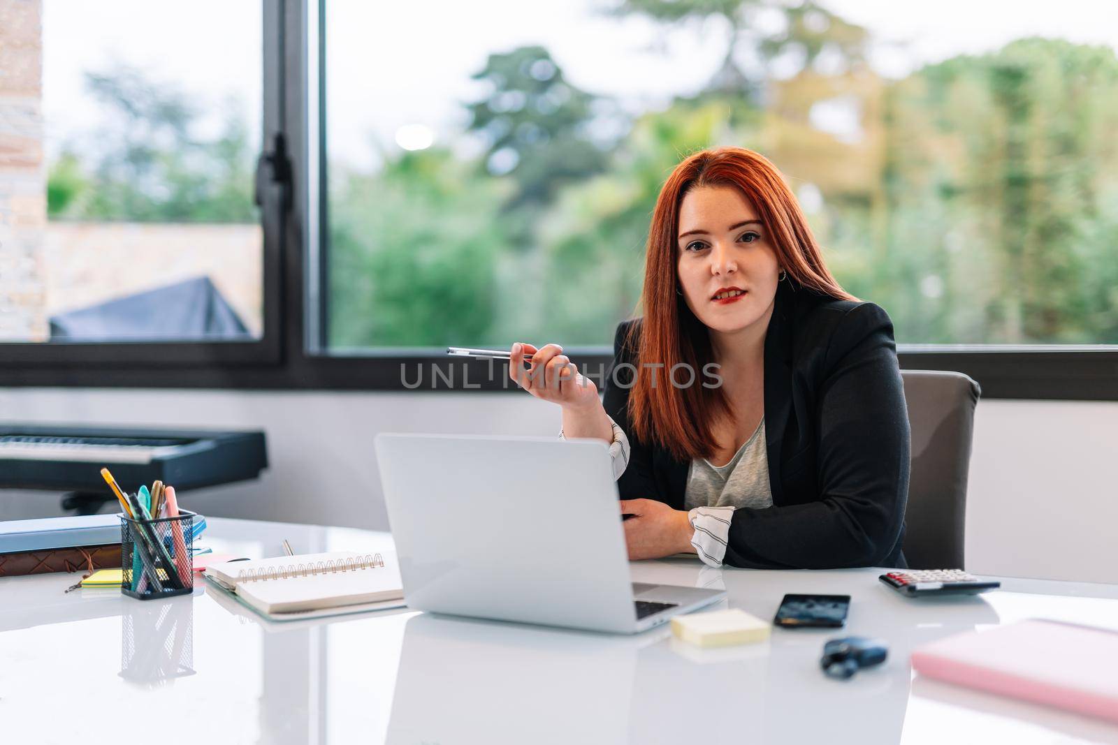 Young red-haired girl teleworking from home in her living room with her white laptop. Brightly lit scene with a large window in the background with natural light. She is wearing a black blazer. Young self-employed businesswoman and entrepreneur working at computer.