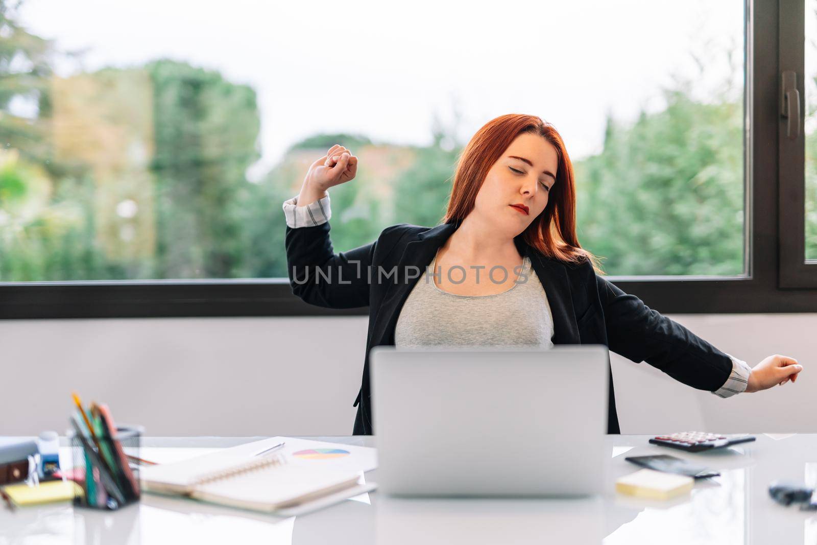 Young red-haired girl in grey T-shirt and black blazer teleworking from home. Enterprising and self-employed businesswoman working in the office or at home. She is stretching her back due to many hours of work at the computer. Large window in background with natural light.