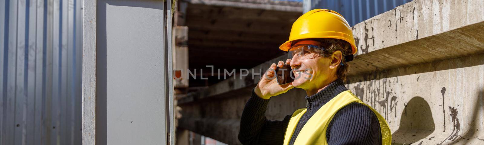 Cheerful matured man in safety helmet having phone conversation and smiling while working at manufacturing plant