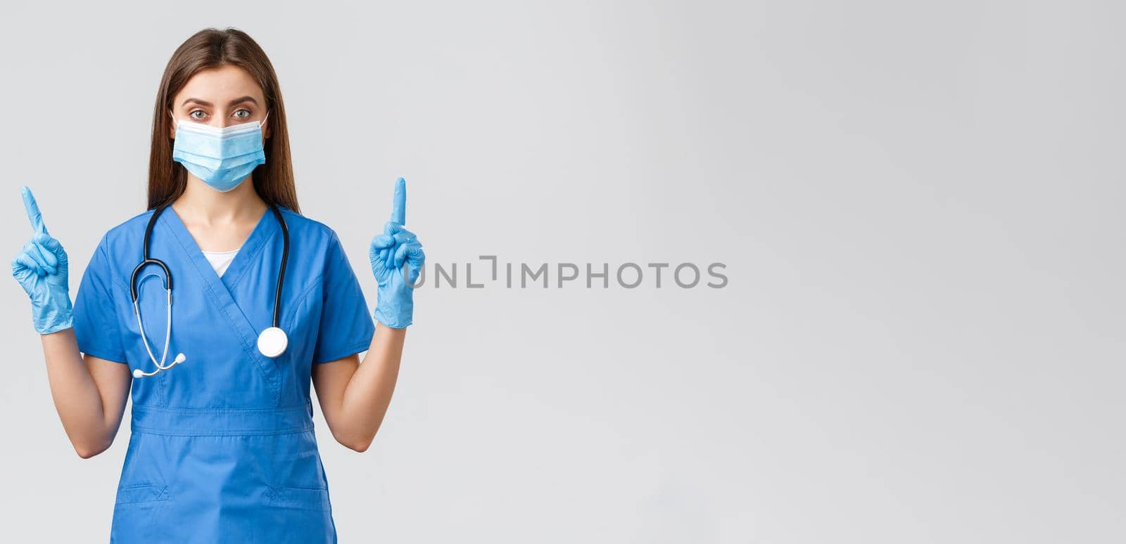 Covid-19, preventing virus, health, healthcare workers concept. Serious and confident female nurse in blue scrubs, medical mask PPE, pointing fingers up, inform patients how prevent corona infection.