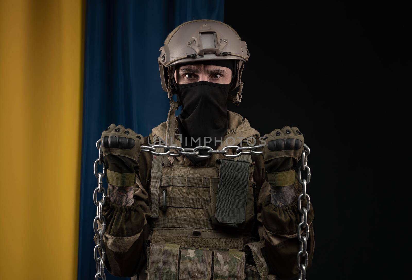 a male soldier in a military uniform and helmet with a weapon on the background of the national flag of Ukraine with a chain as a symbol of slavery