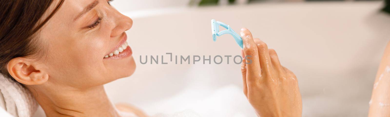 Portrait of lovely smiling young woman holding a razor for shaving her legs and having fun while lying in bathtub by Yaroslav_astakhov