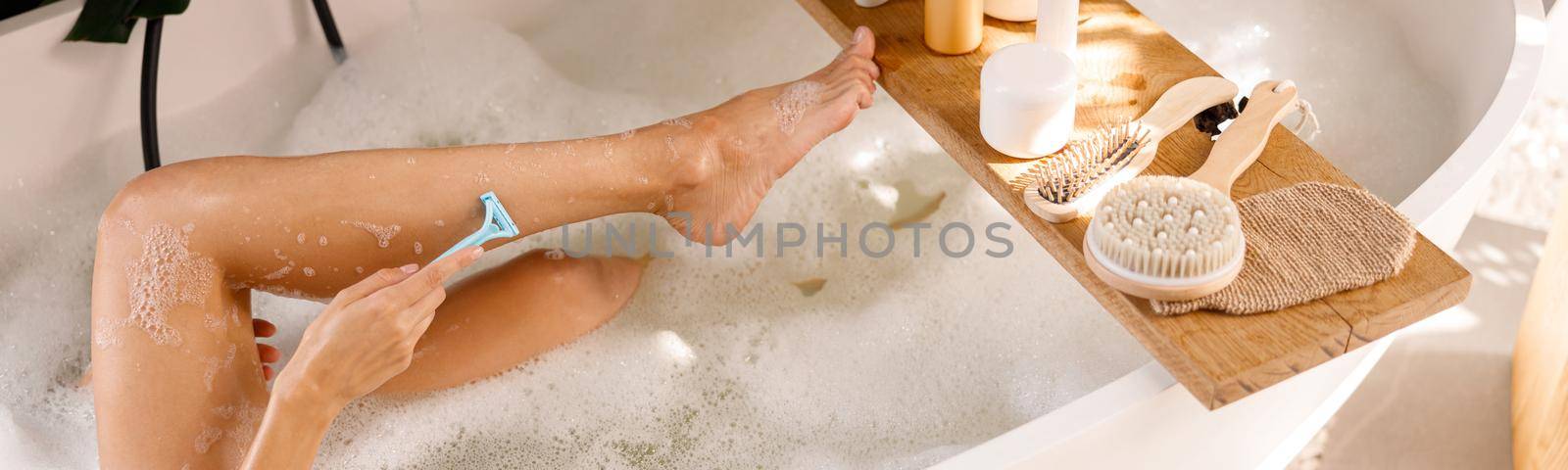 Legs of young woman shaving with disposable shaving razor while bathing. Body care cosmetics on wooden shelf over bathtub. Spa, beauty concept