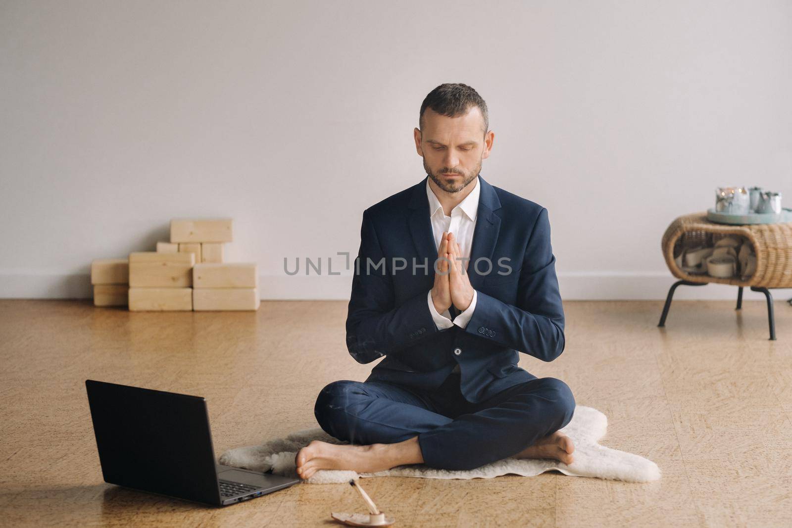 A man in a formal suit meditates while sitting in a fitness room with a laptop.