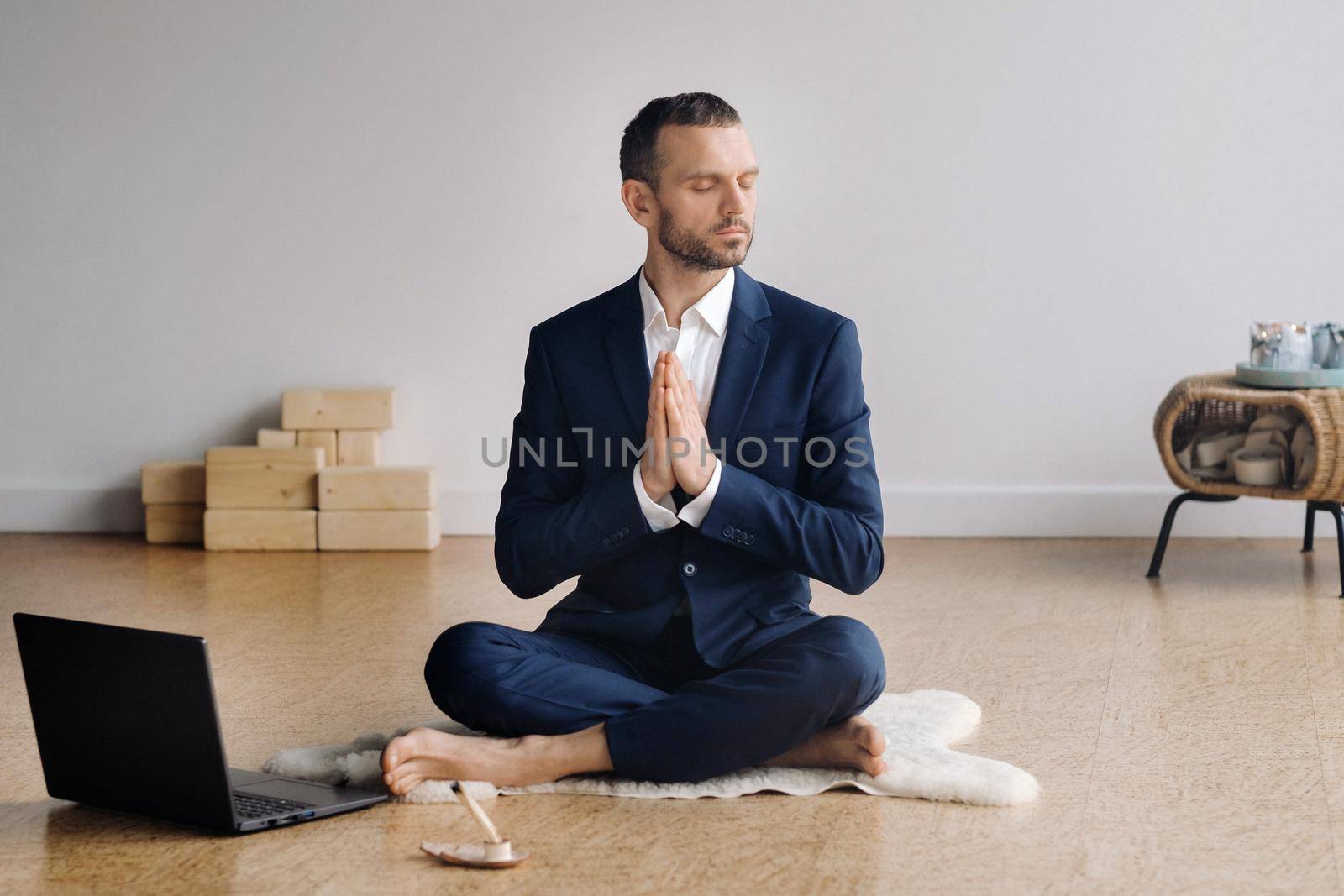 A man in a formal suit meditates while sitting in a fitness room with a laptop.