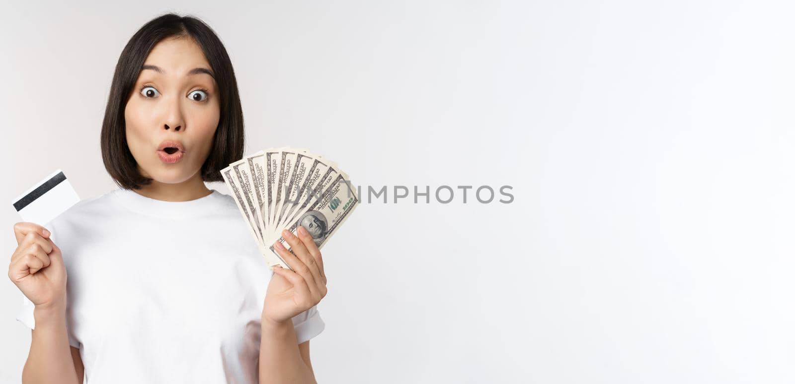 Portrait of asian woman holding money, dollars and credit card, looking impressed and amazed, standing in tshirt over white background.