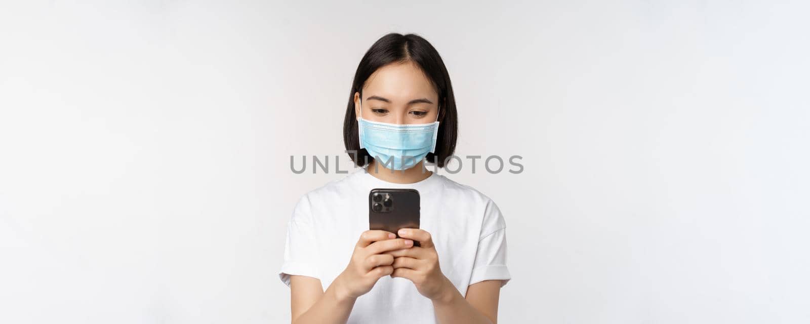 Health, covid and mobile concept. Young asian woman in medical face mask, looking at smartphone screen, using phone app, shopping online, white background.