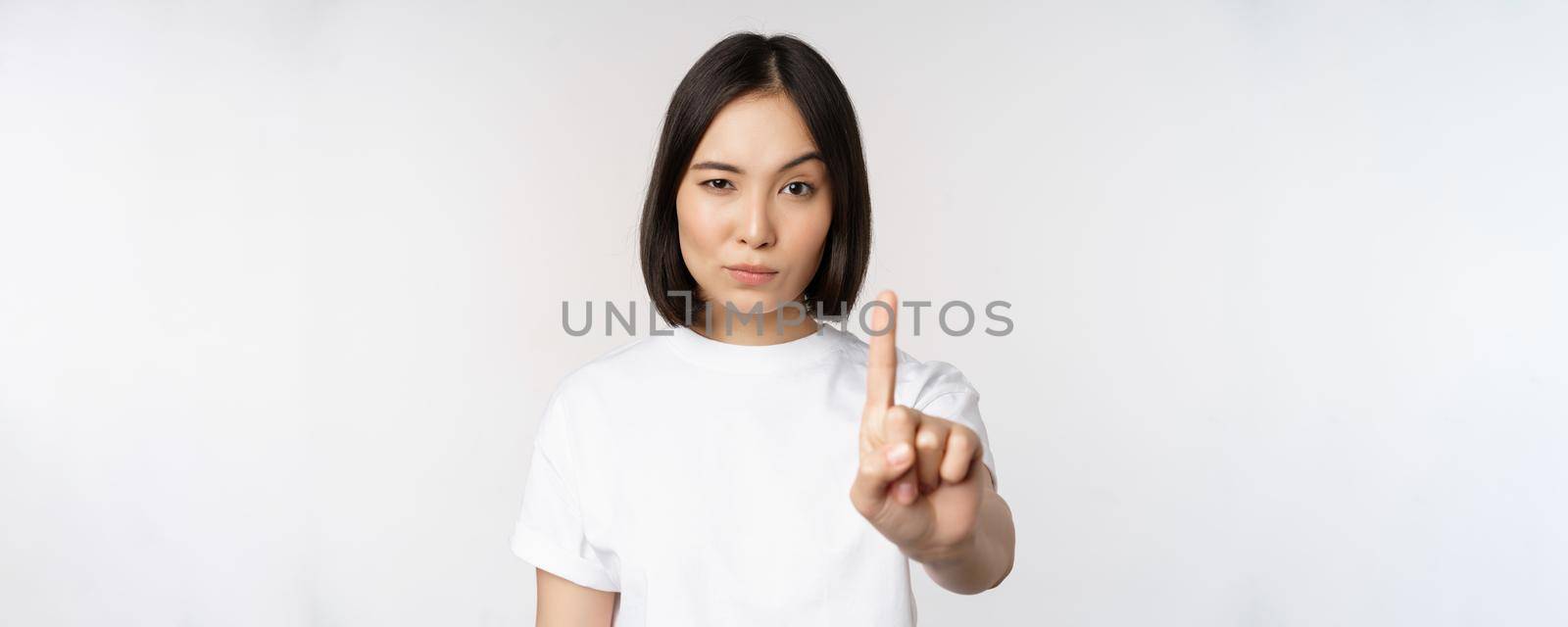 Image of asian girl showing stop, prohibit smth, extend one arm to show forbidding, taboo gesture, standing in tshirt over white background.