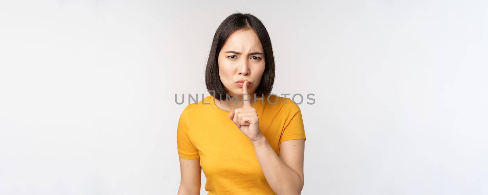 Portrait of angry asian woman shushing, press finger to lips, taboo stop talking sig, looking annoyed, scolding, standing over white background.