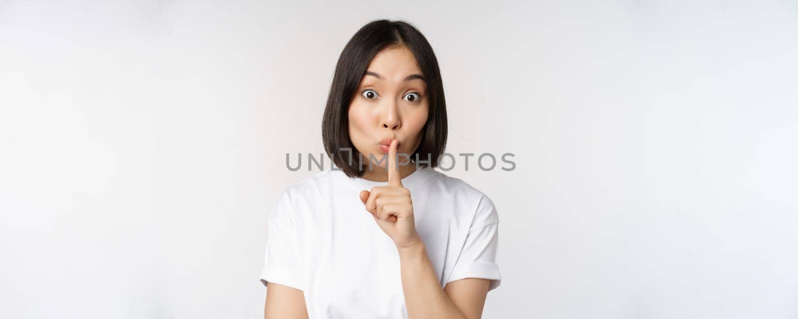 Close up portrait of young beautiful asian girl shushing, has secret, keep quiet silence gesture, press finger to lips, standing in tshirt over white background by Benzoix