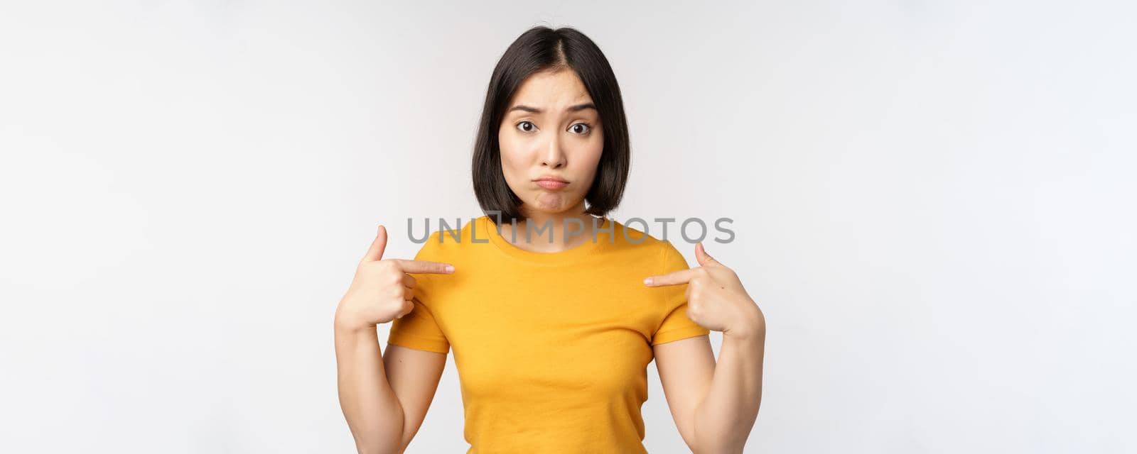 Confused asian woman pointing at herself, looking in disbelief, standing over white background. Copy space