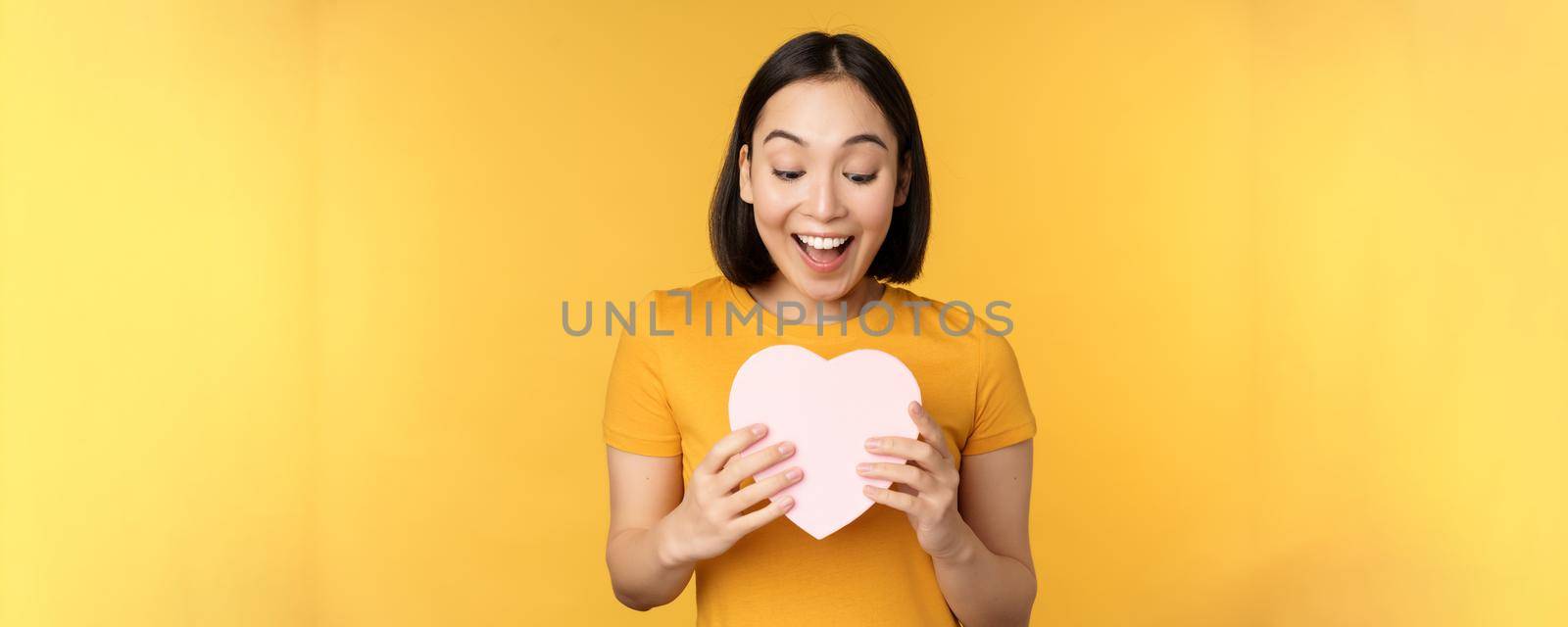 Romance and valentines day. Happy beautiful asian woman holding big heart card and smiling, standing over yellow background by Benzoix